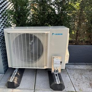 De afbeelding toont een Daikin airco-unit, geplaatst op een vlakke ondergrond en omringd door groen, wat een ideale installatieomgeving suggereert.