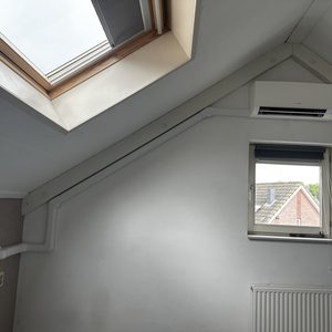 Een wandairconditioner is geïnstalleerd aan de muur, met zichtbare leidingen en een raam in de buurt.