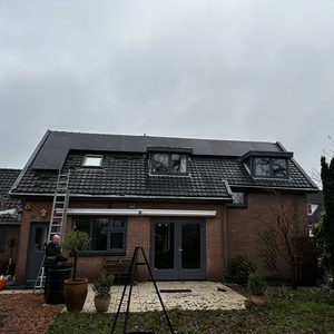 Op het dak van een woning zijn zonnepanelen geïnstalleerd, zichtbaar met een ladder voor toegang. De omgeving is groen en er zijn versterkende elementen zichtbaar.