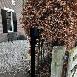 De afbeelding toont een laadpaal voor elektrische voertuigen, geïnstalleerd naast een bomenrij, met een moderne zwarte afwerking.
