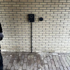 De afbeelding toont een laadpaal voor elektrische voertuigen, bevestigd aan een stenen muur. Kabels lopen van de laadpaal naar de energiebron.