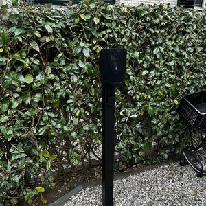 Een laadpaal staat in een tuin, omgeven door groen. Deze installatie is gericht op het opladen van elektrische voertuigen.