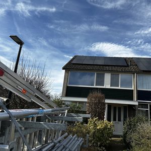 De afbeelding toont een woning met een installatie van zonnepanelen op het dak, omringd door een tuin en gereedschap.