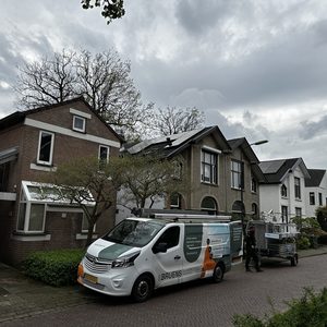 De afbeelding toont een woning met zonnepanelen op het dak, waar installateurs bezig zijn met de installatie. Een bestelwagen is zichtbaar voor de woning.