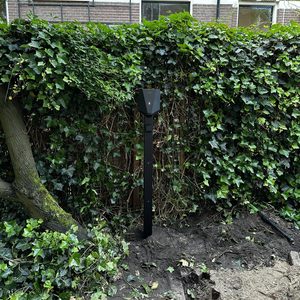 Een laadpaal is geïnstalleerd in de tuin, omringd door groen en een recente graafoperatie.