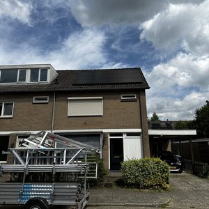 De afbeelding toont een woning met zonnepanelen geïnstalleerd op het dak, samen met een steigerwagen op de voorgrond, wat op een recente installatie wijst.