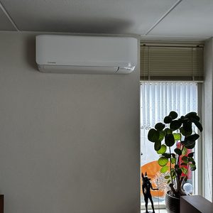 Een wandgemonteerde airconditioningseenheid geïnstalleerd in een woonkamer met een raam en planten op de achtergrond.