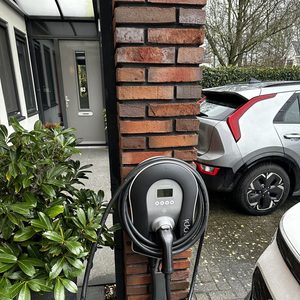 Een laadpaal is geïnstalleerd bij een woning, voorzien van een kabel en stekker voor het opladen van elektrische voertuigen.