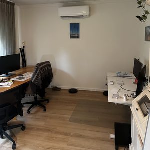 De afbeelding toont een wandgemonteerde airco-installatie in een kantoorruimte, naast twee werkplekken.