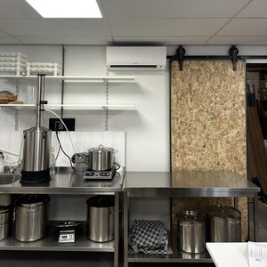 De afbeelding toont een wandmodel airconditioningsysteem geïnstalleerd in een commerciële keukenomgeving, met metaalwerkbladen en verschillende keukenelementen op de achtergrond.