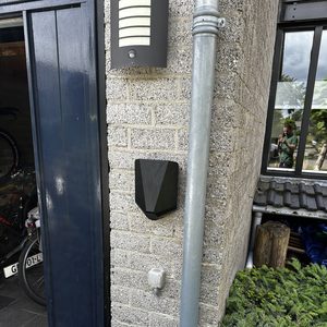 Een laadpaal is geïnstalleerd aan de muur, dichtbij een deur en omgeven door planten en een metalen buis.