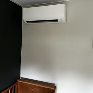 Een wandairconditioner van het merk Daikin is geïnstalleerd in een ruimte. Het apparaat is geplaatst boven een houten bedje.