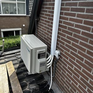 De afbeelding toont een buitenunit van een airconditioning installatie, gemonteerd op een dak met zichtbare verbindingen naar de woning.