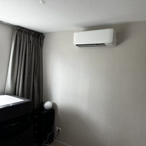 De afbeelding toont een wandairconditioner geïnstalleerd op een muur in een kamer, met een bed en een nachtkastje in de buurt.