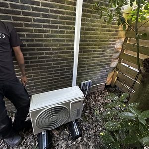 De afbeelding toont een buitenunit van een airconditioning installatie, geplaatst op een fundering met aansluitingen aan de wand.