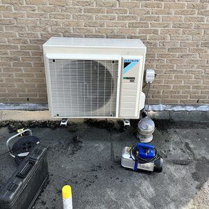 De afbeelding toont een buitenunit van een Daikin airconditioning systeem, geïnstalleerd tegen een bakstenen muur, met verschillende gereedschappen en accessoires op de vloer.