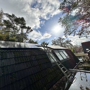 Een installatie van zonnepanelen op een hellend dak, met ladders en bomen op de achtergrond. De panelen vangen zonlicht voor energieproductie.