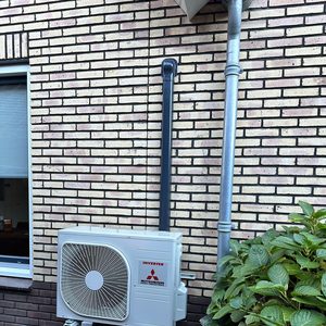 De installatie toont een buitenunit van een airconditioning van Mitsubishi, bevestigd aan een stenen muur met zichtbare leidingen.