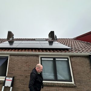 De afbeelding toont een woning met zonnepanelen geïnstalleerd op het schuine dak. Een man loopt ernaast, waarschijnlijk betrokken bij de installatie.