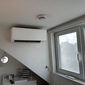 Een wandmontage airconditioner die efficiënt koele lucht verspreidt in een ruimte. Installatie bevindt zich onder een schuin plafond vlakbij een raam.