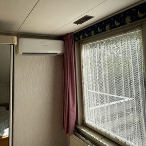Een wandairconditioner is geïnstalleerd in een kamer, zichtbaar boven een verwarmingselement en een raam met gordijnen.