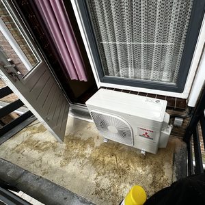 De afbeelding toont een airco-installatie buiten op een balkon, met zicht op de open deur naar binnen.