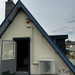 Externe airconditioninginstallatie bevestigd aan de muur van een woning, met leidingen voor koelmiddel en elektriciteit zichtbaar.