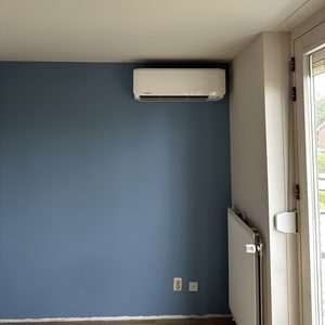 Een wandunit voor airconditioning is geïnstalleerd op een blauwe muur, met zicht op een radiator en een deur naar buiten.