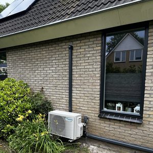 De afbeelding toont een wandairco-installatie met een buitenunit, geplaatst naast een woning met zonnepanelen op het dak.