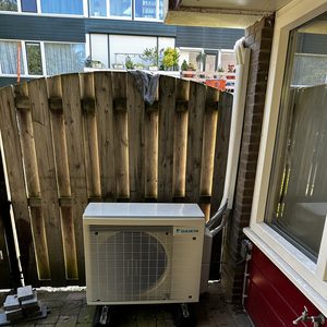 De foto toont een buitenunit van een Daikin airconditioning, geïnstalleerd op een stenen ondergrond in een tuinomgeving.