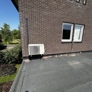 De afbeelding toont een buitenunit van een airconditioning installatie aan de muur van een huis, met een zonnige omgeving op de achtergrond.