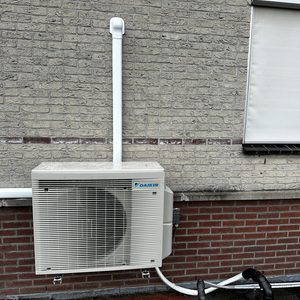 De afbeelding toont een buitenunit van een airconditioningssysteem, geïnstalleerd aan de muur met bijbehorende leidingen.