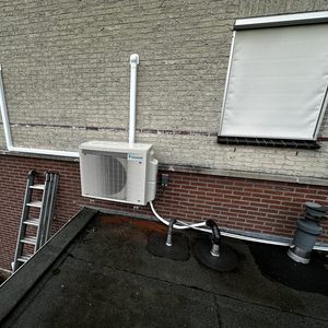 Een buitenunit van een airconditioning, geïnstalleerd aan de muur van een gebouw, inclusief leidingen en een ladder nabij.