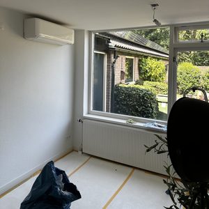 Een wand-airconditioner is geïnstalleerd in een lichte kamer met uitzicht op een tuin.