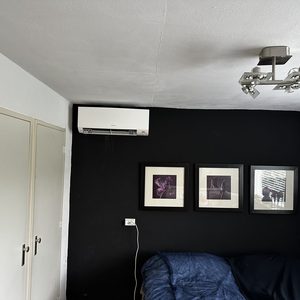 Een wandairconditioner is gemonteerd in een donkere kamer, met kunst aan de muur en een bed op de voorgrond.