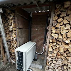Een airco-unit is geïnstalleerd in een schuurachtige ruimte, omringd door houtstapels. De installatie heeft een nette afwerking met leidingen naar de buitenzijde.