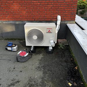 Een wandunit voor airconditioning geïnstalleerd aan een bakstenen muur, met leidingen en accessoires op de vloer ernaast.