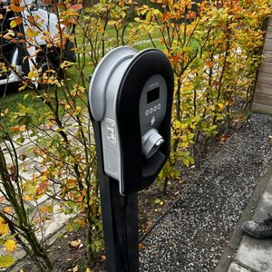 De afbeelding toont een laadpaal voor elektrische voertuigen, geplaatst in een tuin met groene elementen op de achtergrond.