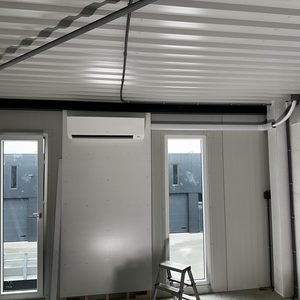 Een airconditioninginstallatie hangt aan de muur tussen twee ramen in een kamersituatie, met zichtbare leidingen.