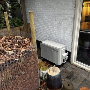 De afbeelding toont een buitenunit van een airconditioningsysteem, geplaatst naast een muur en omringd door bladeren en andere tuinmaterialen.