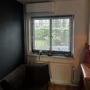 Een wandmodel airconditioner geïnstalleerd naast een raam met jaloezieën, in een woonkamer setting.