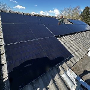 De afbeelding toont een installatie van zonnepanelen op een schuin dak, met een installateur bezig bij de panelen.