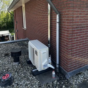 De afbeelding toont een buitenunit van een airconditioning installatie, geplaatst op een betegeld oppervlak tegen een bakstenen muur.