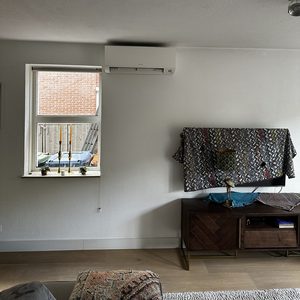 Een wand-airconditioningseen in een woonkamer, geïnstalleerd boven een venster voor optimale luchtcirculatie.