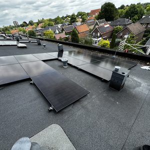 De foto toont een installatie van zonnepanelen op een vlakke dakoppervlakte, met een overzicht van meerdere panelen en omgevingsarchitectuur.