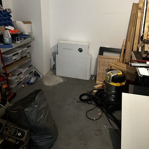 De foto toont een thuisbatterij installatie in een opslagruimte, met afval en gereedschap in de omgeving.
