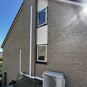 Een buitenunit van een airco-installatie met bijbehorende leidingen, geplaatst tegen een stenen muur.