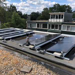 De foto toont een onvoltooide installatie van zonnepanelen op een plat dak, met montagemateriaal en leidingen zichtbaar.